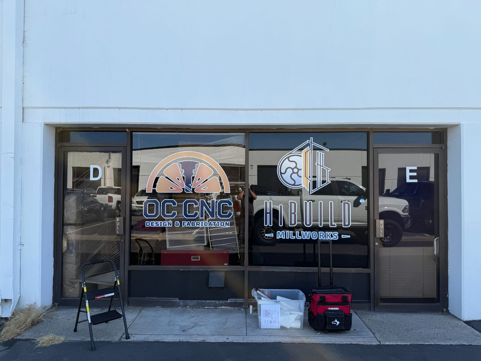 Industrial unit storefront glass with two tenants' printed vinyl logos applied to the windows: 'OC CNC Design & Fabrication' and 'HiBuild Millworks', beside doors labeled D and E