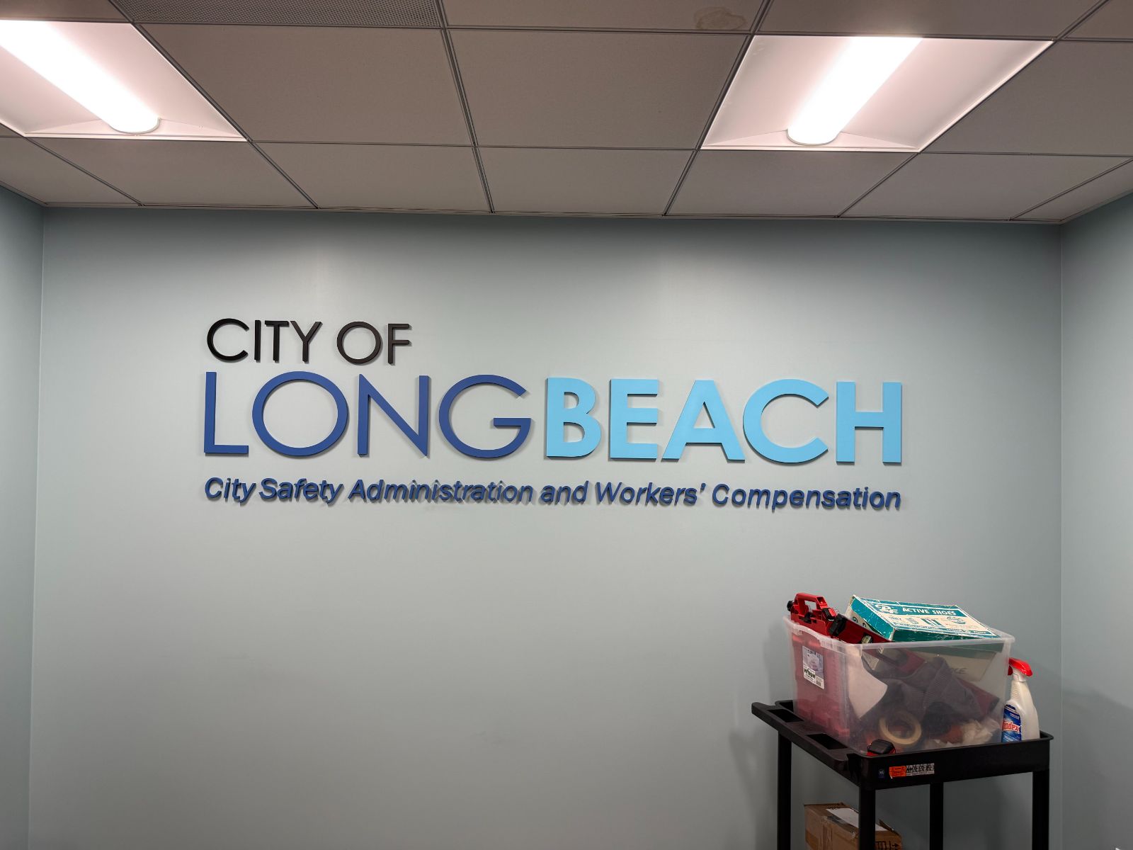 Interior wall with 'City of Long Beach' in dimensional letters: black, blue and light-blue faces above a line reading 'City Safety Administration and Workers' Compensation,' flush-mounted on a pale wall