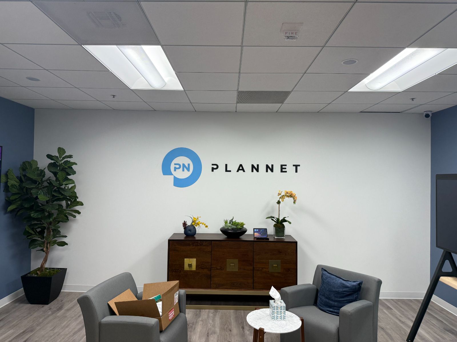 Reception wall in an office lobby with the PLANNET logo: a blue speech-bubble 'PN' mark beside black flat-cut letters spelling PLANNET, above a wood credenza