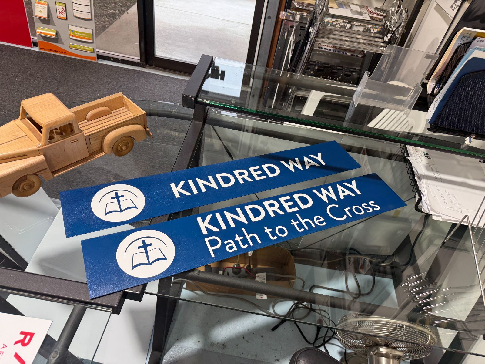 Two blue directional signs on a shop counter reading 'Kindred Way' and 'Kindred Way: Path to the Cross,' each with a white circular open-book-and-cross emblem, printed on aluminum blanks