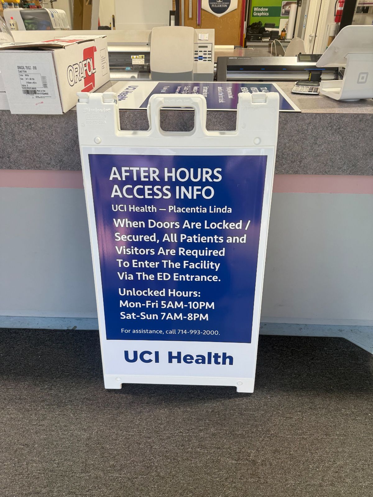 White plastic A-frame sidewalk sign with a blue printed 'UCI Health: After Hours Access Info' panel listing entry instructions and unlocked hours, standing at the Hi Sign shop counter