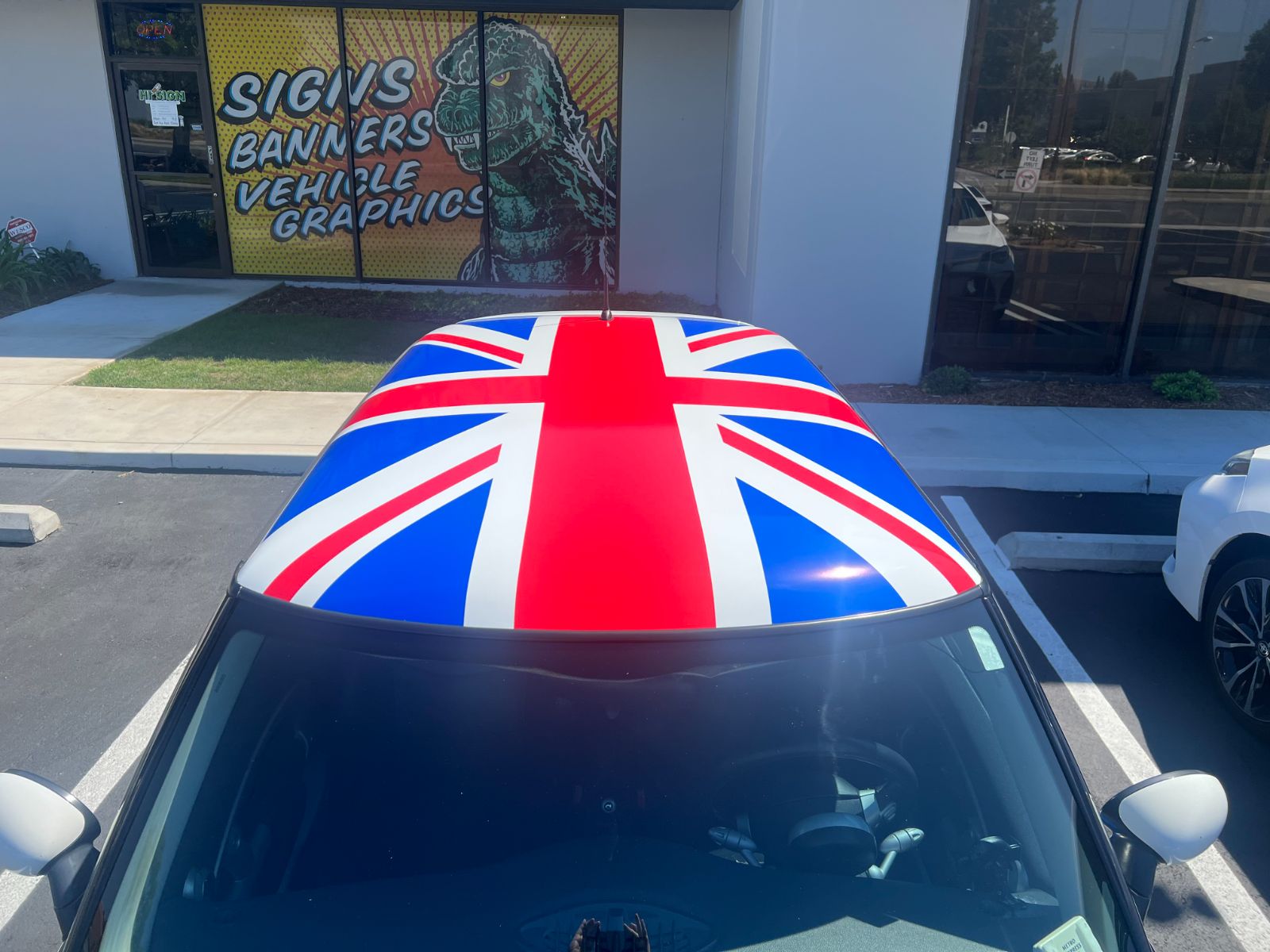 Roof of a MINI Cooper wrapped in a printed Union Jack flag graphic, parked in front of the Hi Sign shop window that reads 'Signs, Banners, Vehicle Graphics'