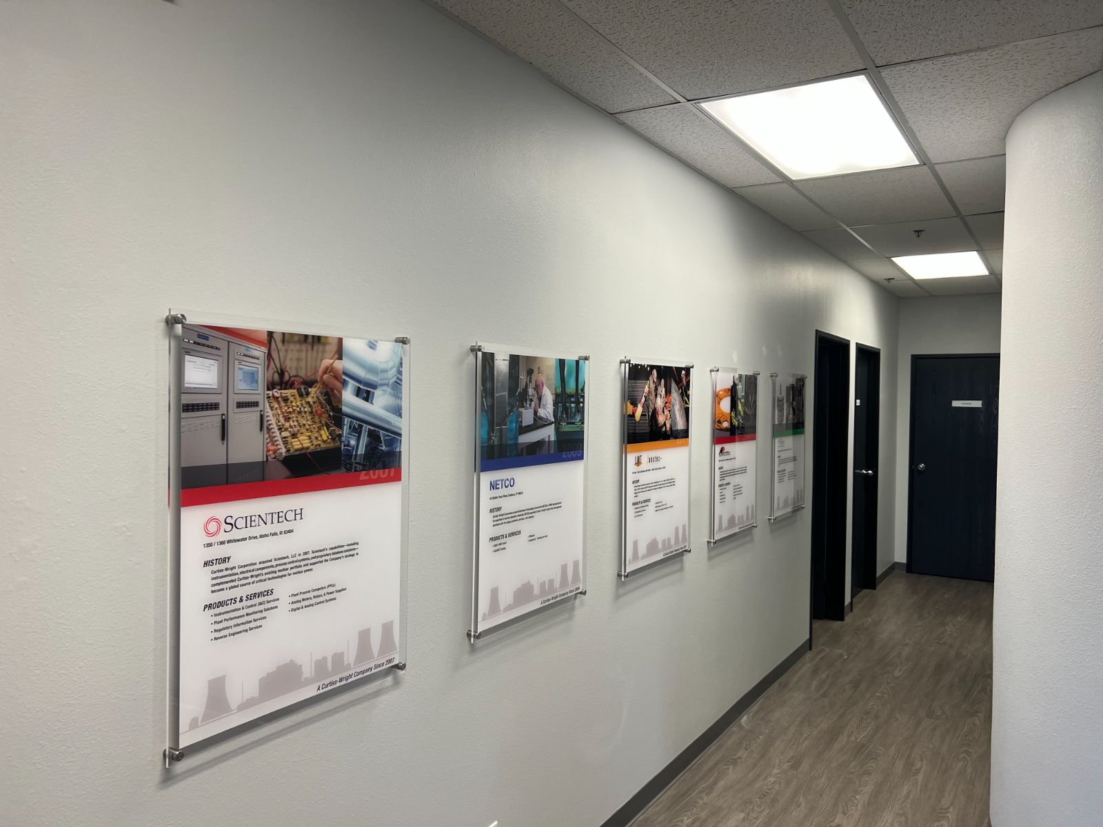 Corporate history wall in an office corridor: a row of printed company milestone panels (Scientech, NETCO, and other Curtiss-Wright brands) mounted in clear acrylic standoff frames along a white interior hallway