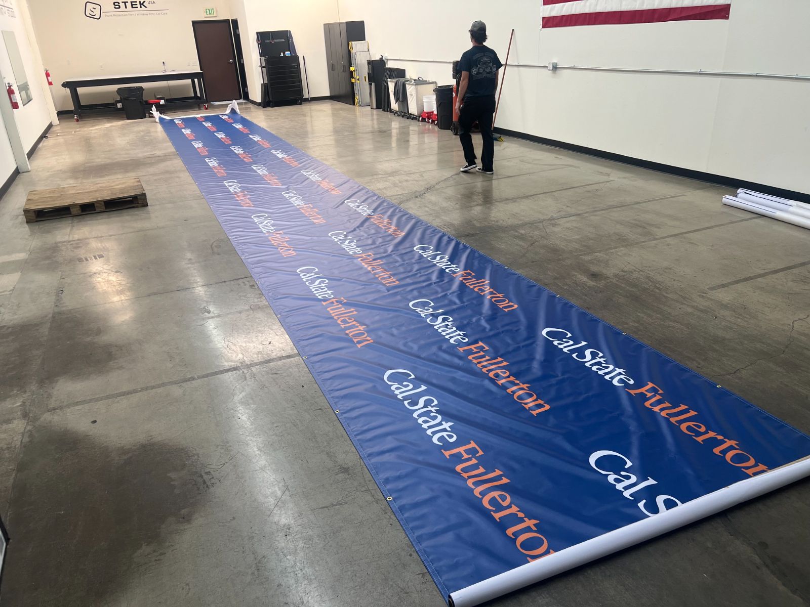 A very long Cal State Fullerton step-and-repeat banner unrolled across a shop floor: blue printed vinyl with the university wordmark repeated in white and orange, grommeted along the edges