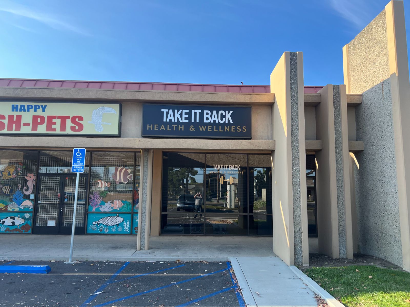 Storefront fascia sign cabinet reading 'Take It Back: Health & Wellness' in white and gold on a black face, mounted above the entrance of a strip-mall unit next to a pet store