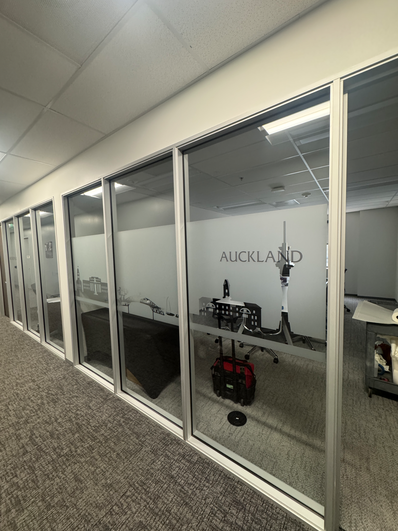 Corridor of glass-walled meeting rooms, each with a frosted privacy band carrying a cut-vinyl city skyline and room name, including AUCKLAND with its harbour bridge and tower