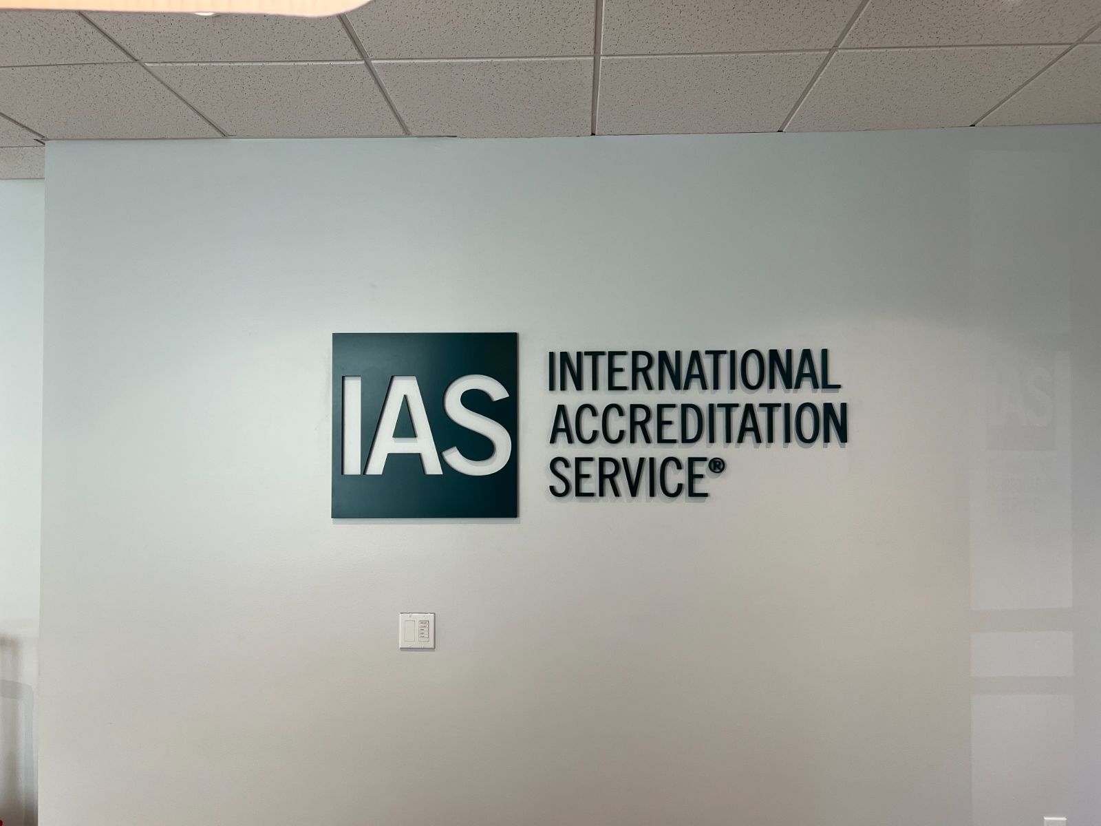 IAS (International Accreditation Service) dimensional lobby logo: dark teal ACM backer panel with raised white cast acrylic letterforms, flanked by dimensional near-black painted letters spelling 'International Accreditation Service,' standoff-mounted on a white interior office wall