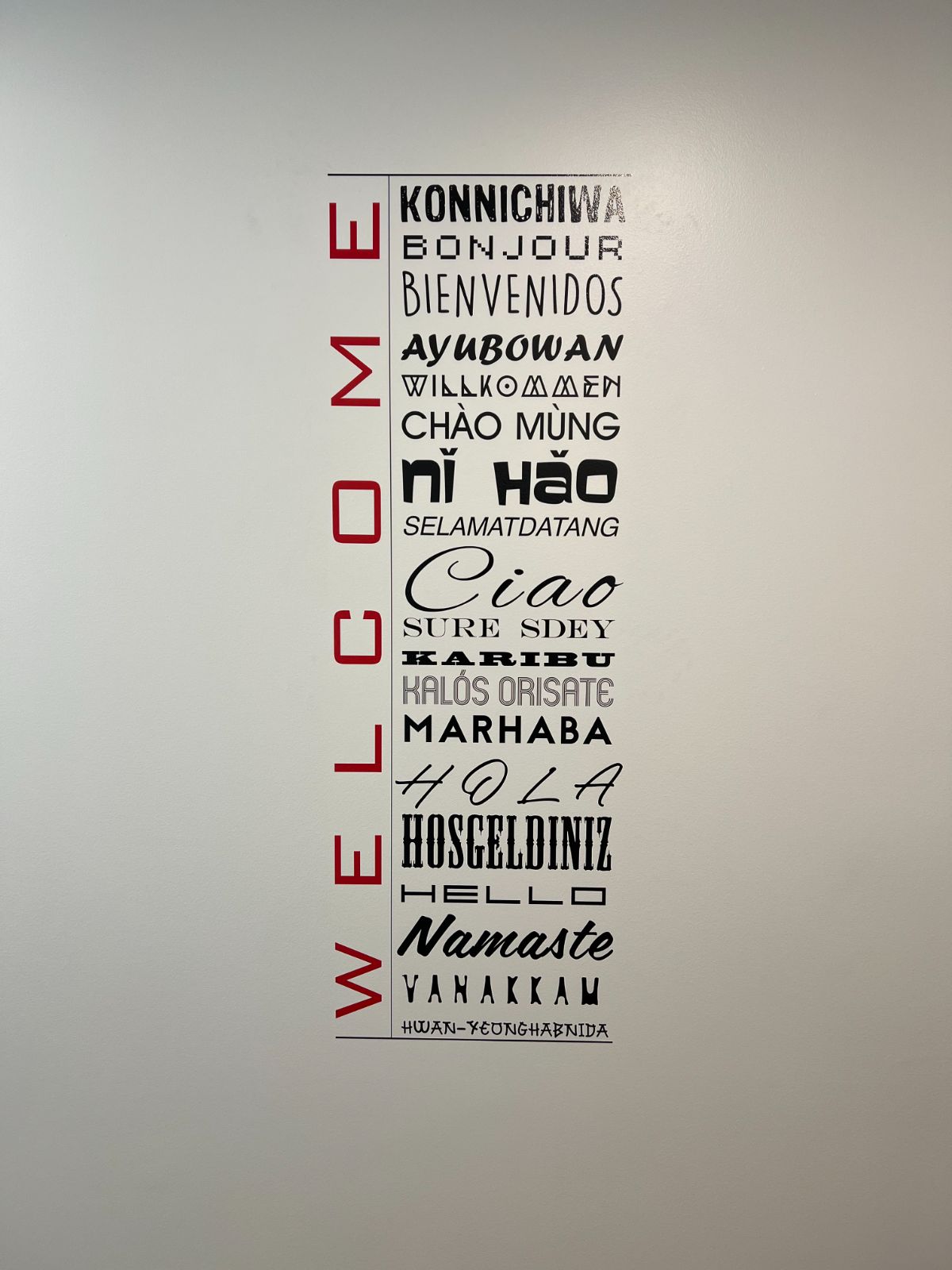Multi-language welcome wall graphic: vertical red cut vinyl 'WELCOME' letters beside a cut vinyl panel listing greetings in 18 languages including Konnichiwa, Bonjour, Bienvenidos, Nǐ hǎo, and Namaste, applied to a white interior wall