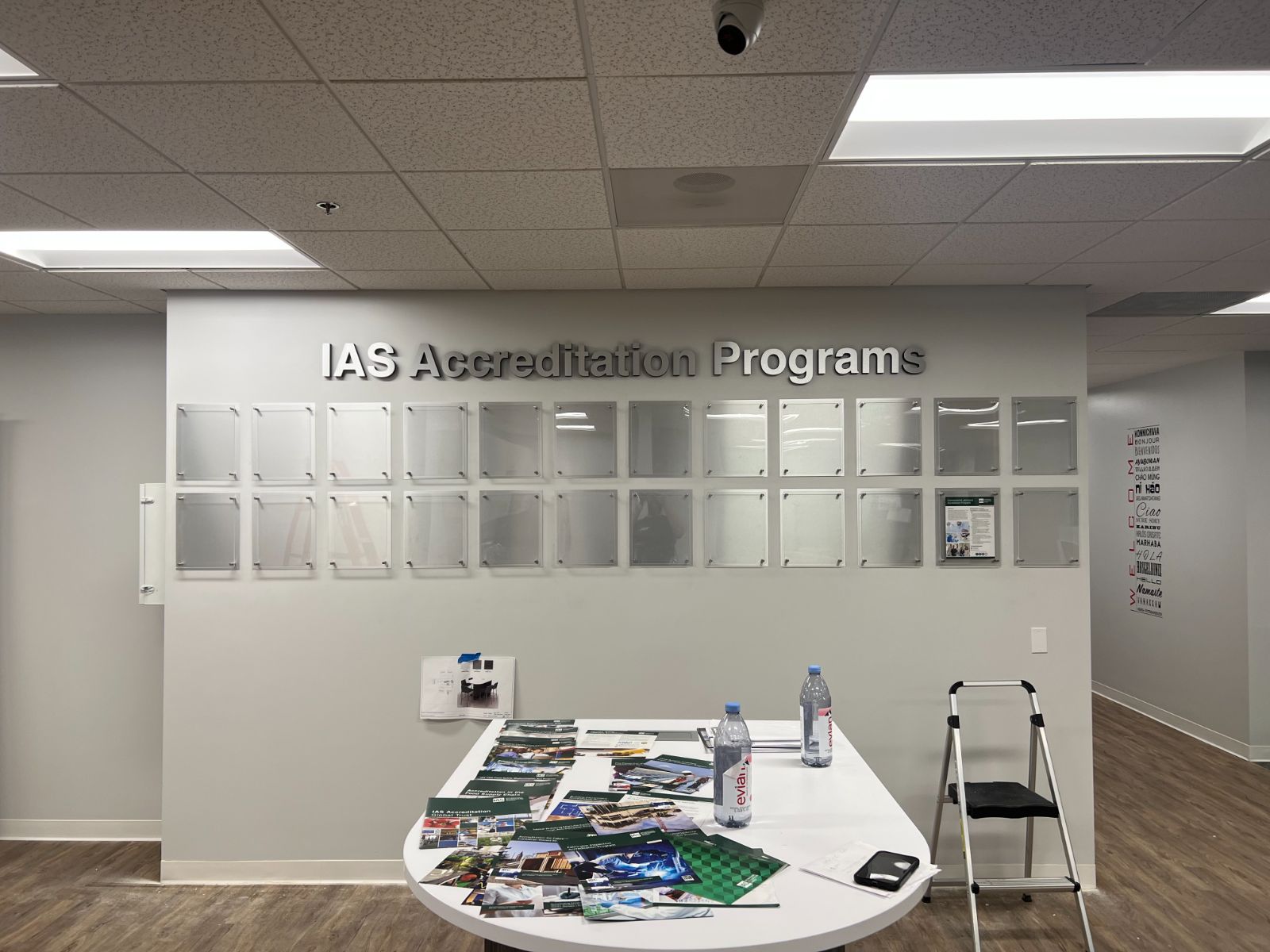 IAS Accreditation Programs corporate feature wall: large brushed aluminum dimensional letters as headline above a 4×5 grid of clear acrylic standoff-mounted document display frames on a white interior office wall