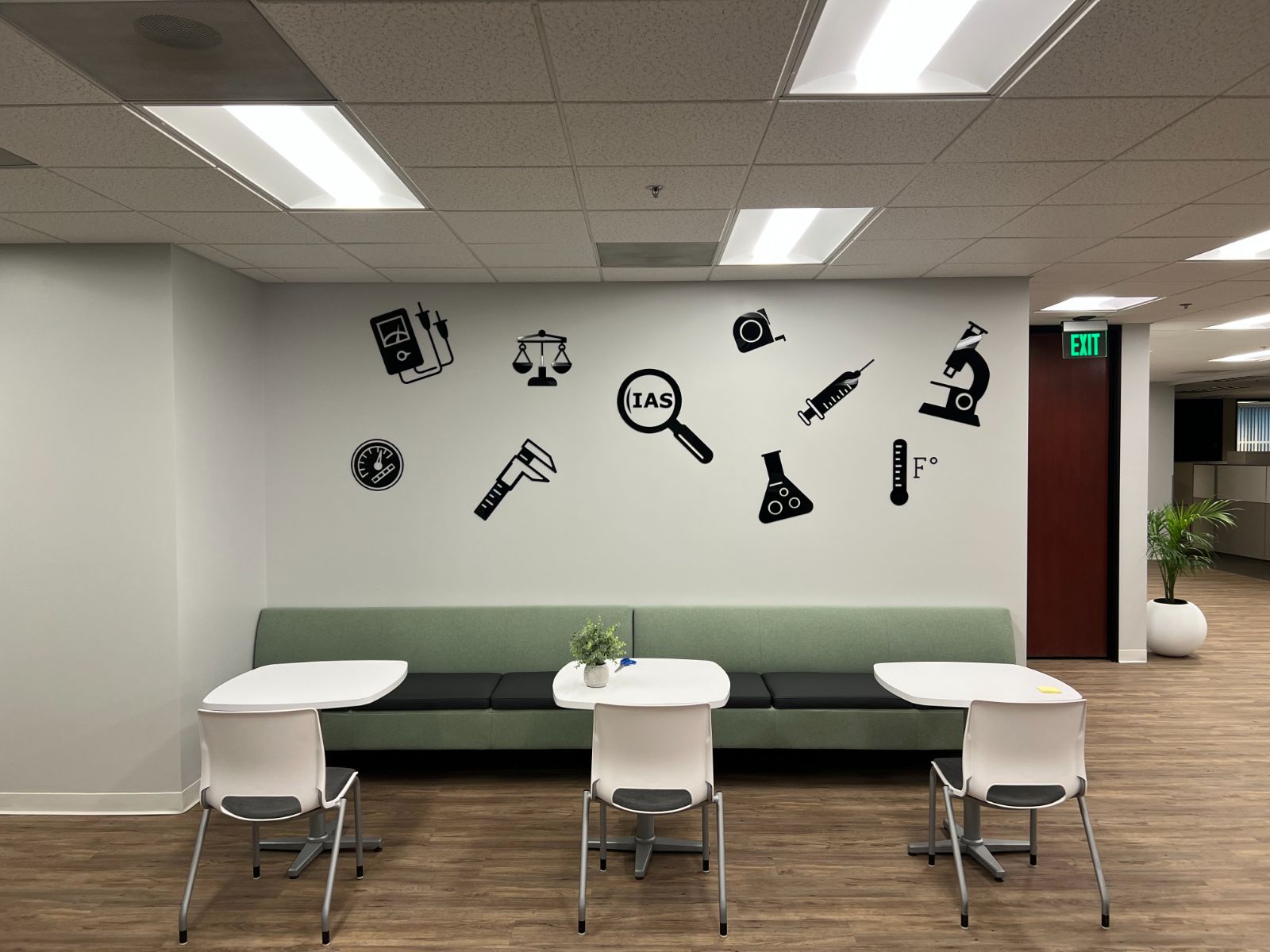 Office break area with a scatter of black cut-vinyl instrument icons on the wall: a multimeter, scale, calipers, magnifier with 'IAS', tape measure, syringe, flask, thermometer and microscope, above a green bench