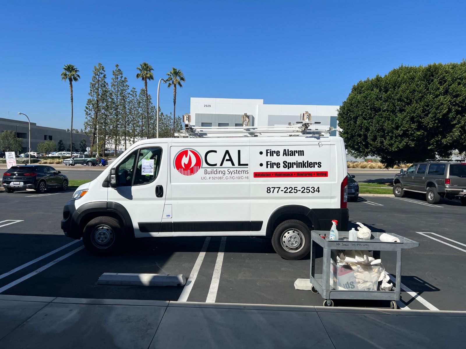 Passenger side of the same white CAL Building Systems van in a parking lot: vinyl logo, 'Fire Alarm / Fire Sprinklers' lettering and phone number on the body panel