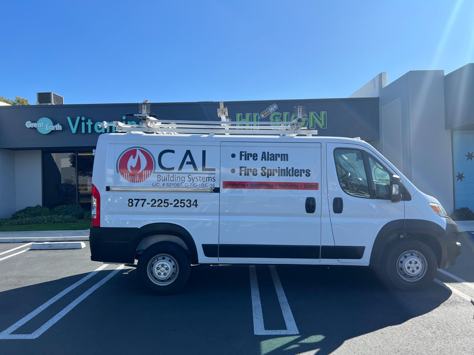 Driver side of a white CAL Building Systems cargo van parked outside the Hi Sign shop: cut vinyl fire-flame logo, 'Fire Alarm / Fire Sprinklers' lettering, phone number, and license numbers applied to the body panel