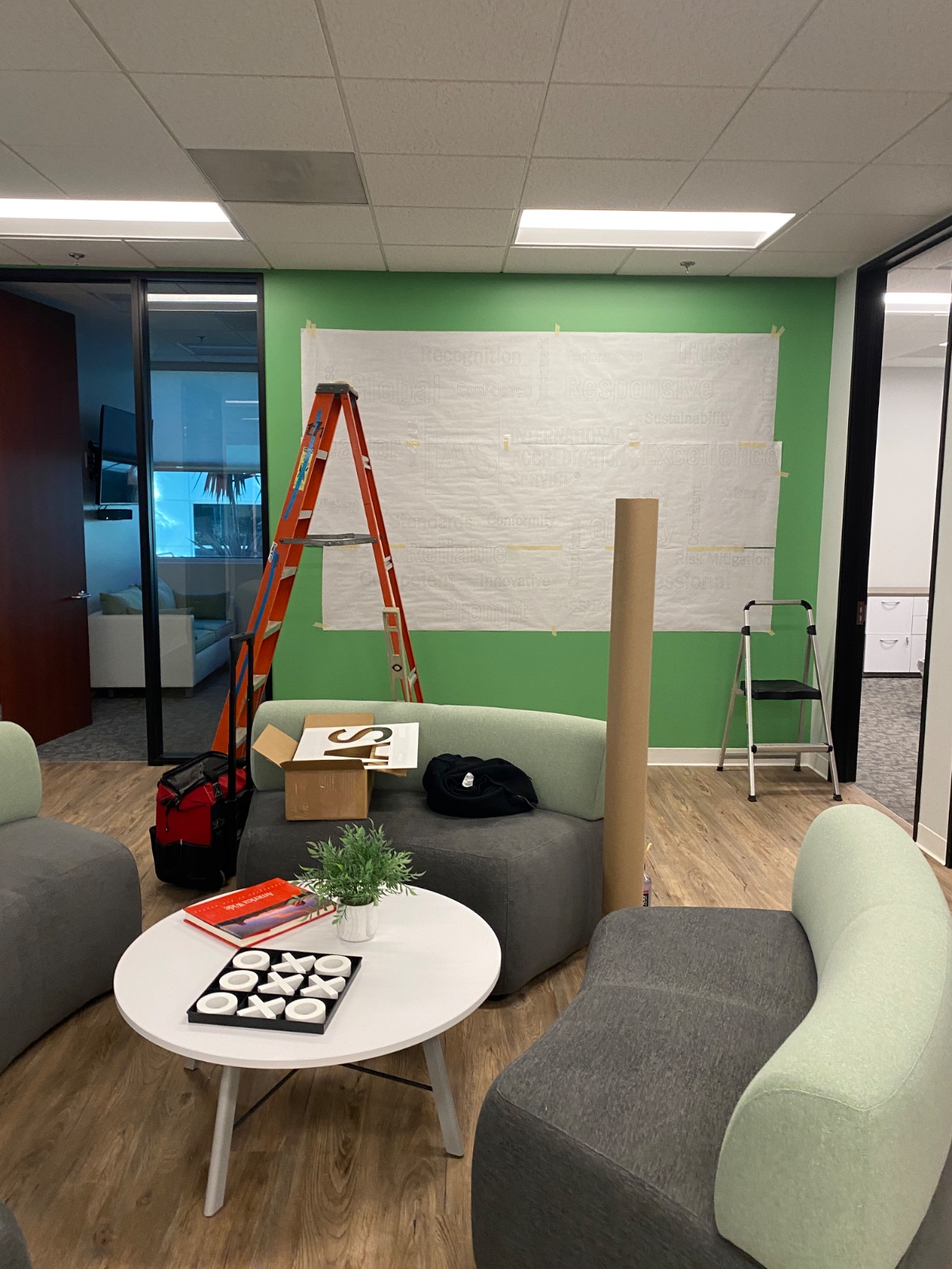Office lounge with a full-size paper layout template taped to a green wall, marking the position of a word-cloud wall graphic before the vinyl goes up