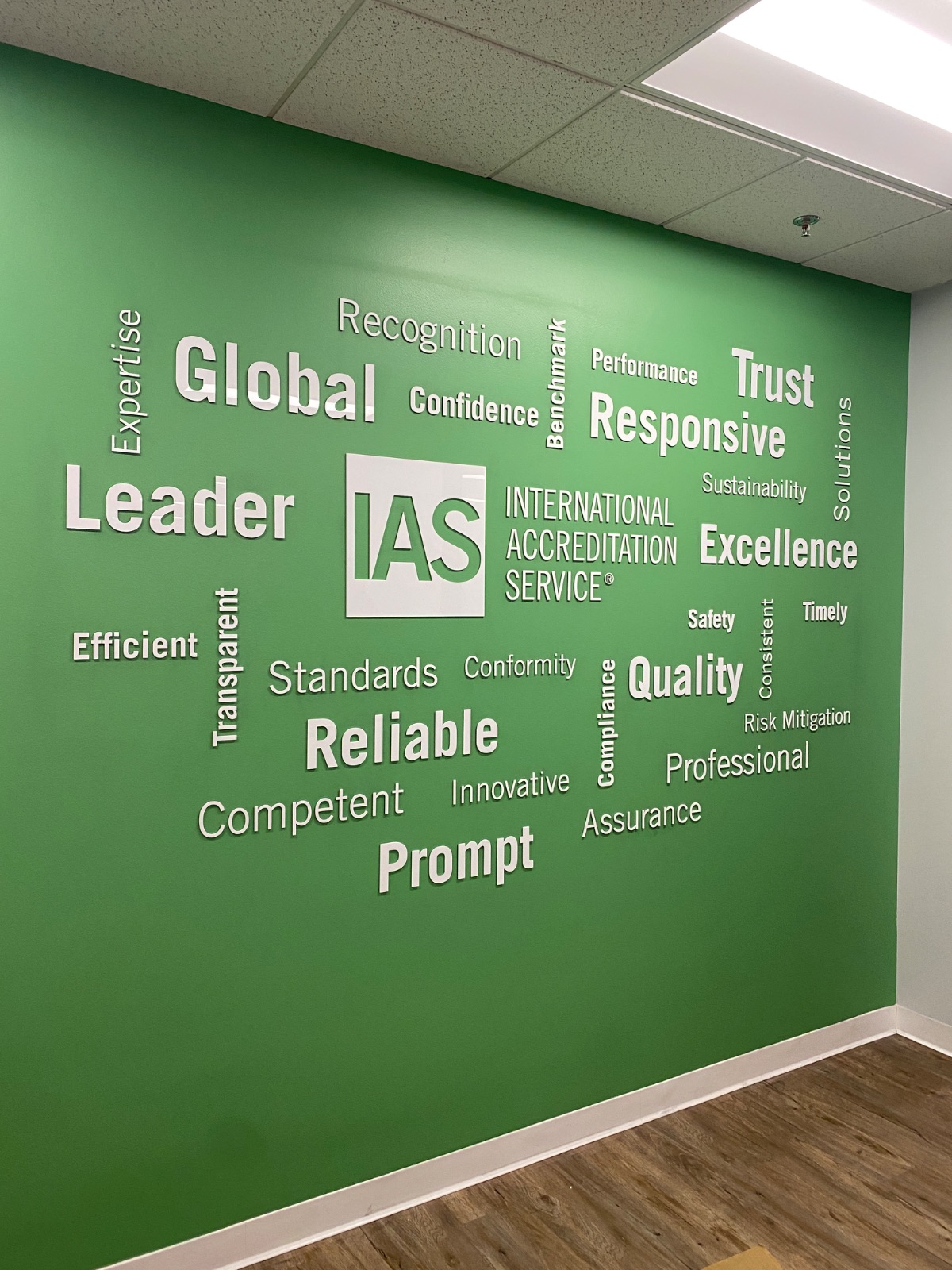 Green office wall covered in a cut-vinyl word cloud of white and grey terms (Global, Leader, Trust, Responsive, Excellence, Reliable, Prompt) around the IAS logo