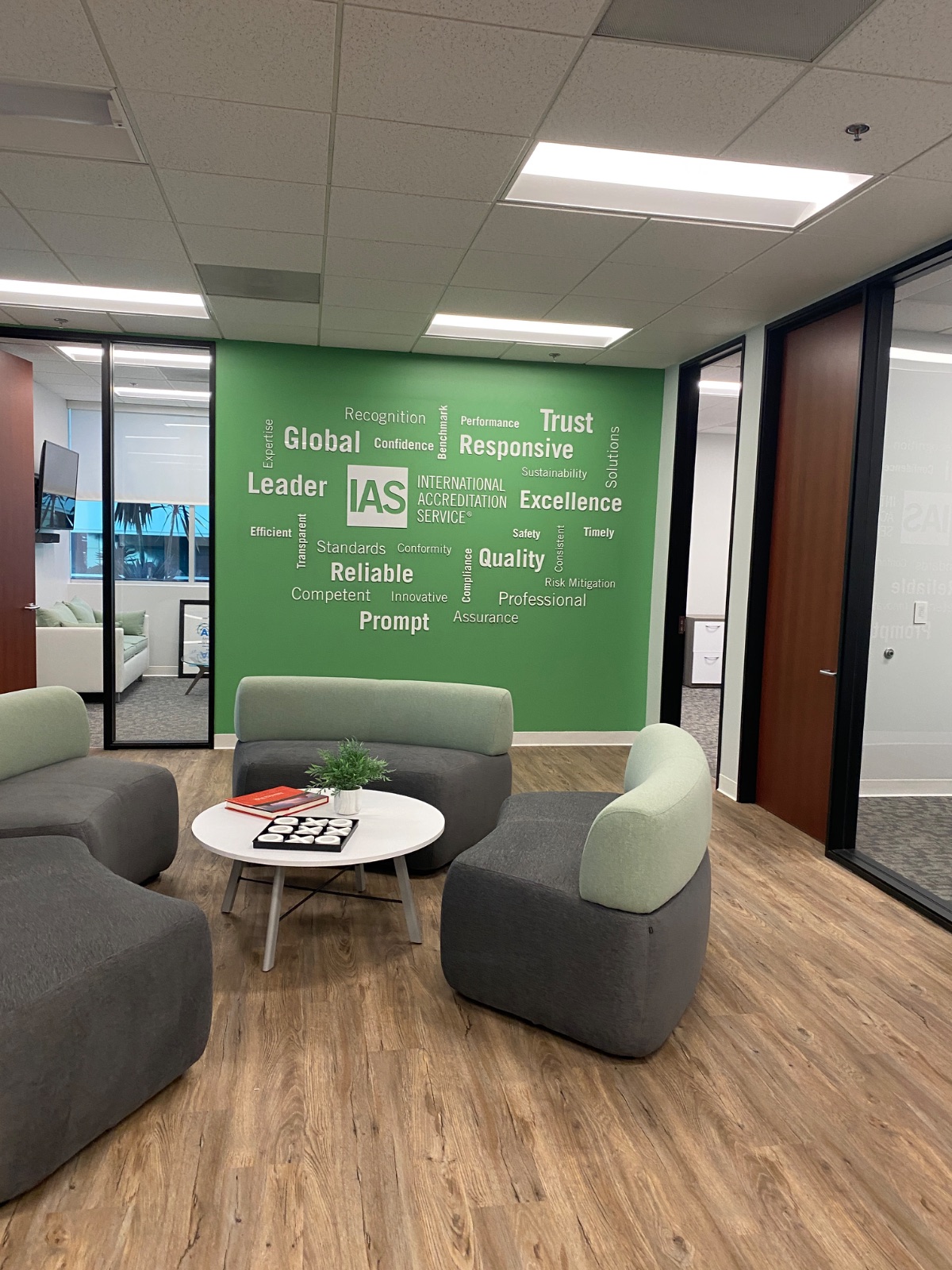 IAS (International Accreditation Service) lobby feature wall: dimensional white IAS logo panel standoff-mounted at the center of a green word-cloud wall graphic, installed by Hi Sign, Brea CA