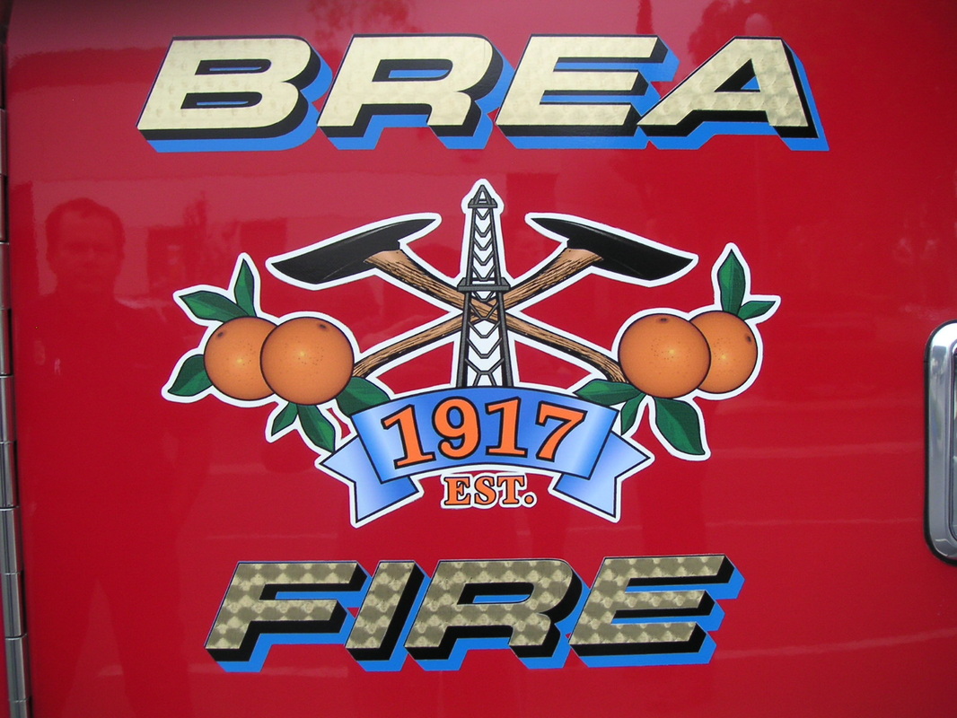 Door of a red fire engine with 'BREA' and 'FIRE' in gold-leaf-look dimensional lettering above and below a printed Brea Fire crest of crossed axes, oranges, and an oil derrick with an EST. 1917 banner