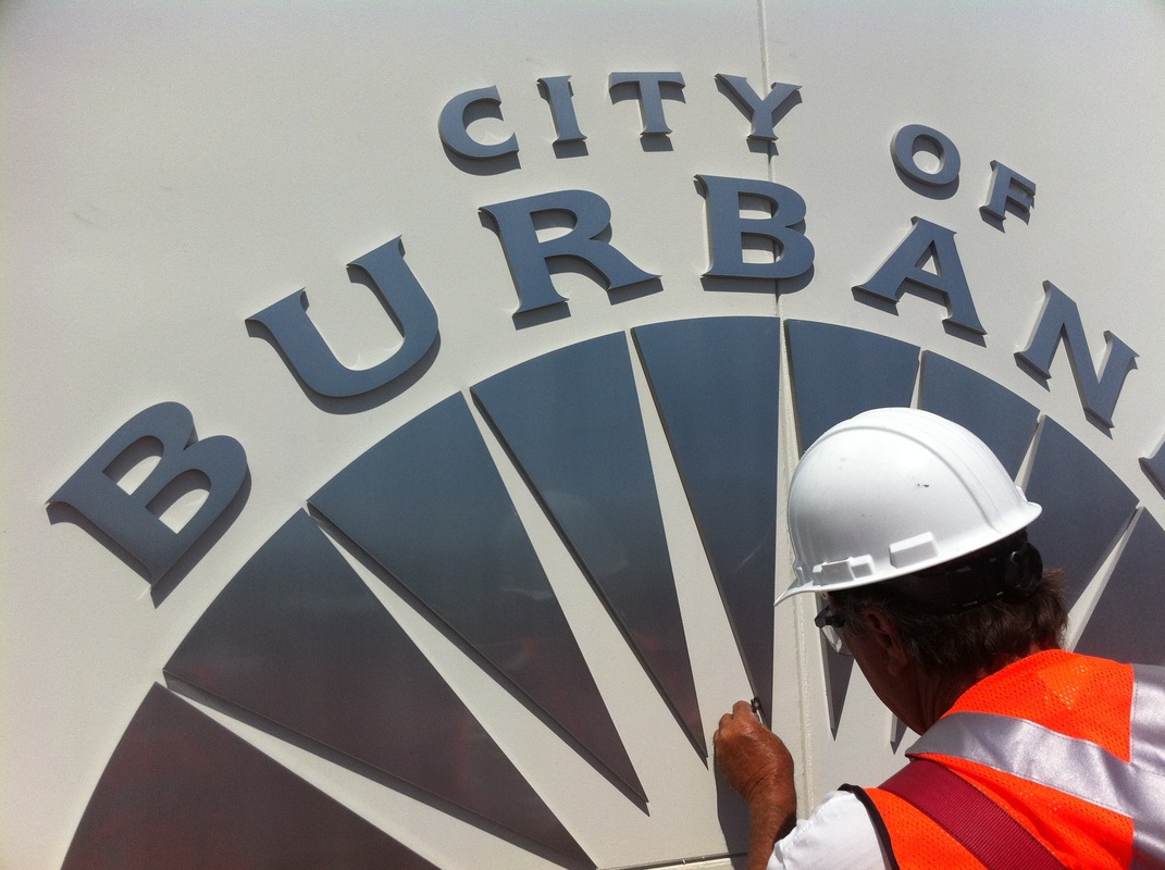 Installer in a hard hat and safety vest setting gray dimensional letters spelling CITY OF BURBANK around a sunburst seal on a white exterior panel