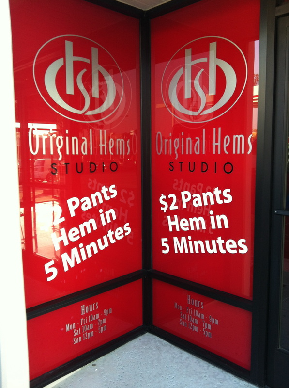 Storefront entry with red vinyl window graphics for Original Hems Studio: a silver monogram, business name, white offer copy reading '$2 Pants Hem in 5 Minutes,' and store hours across two glass panels