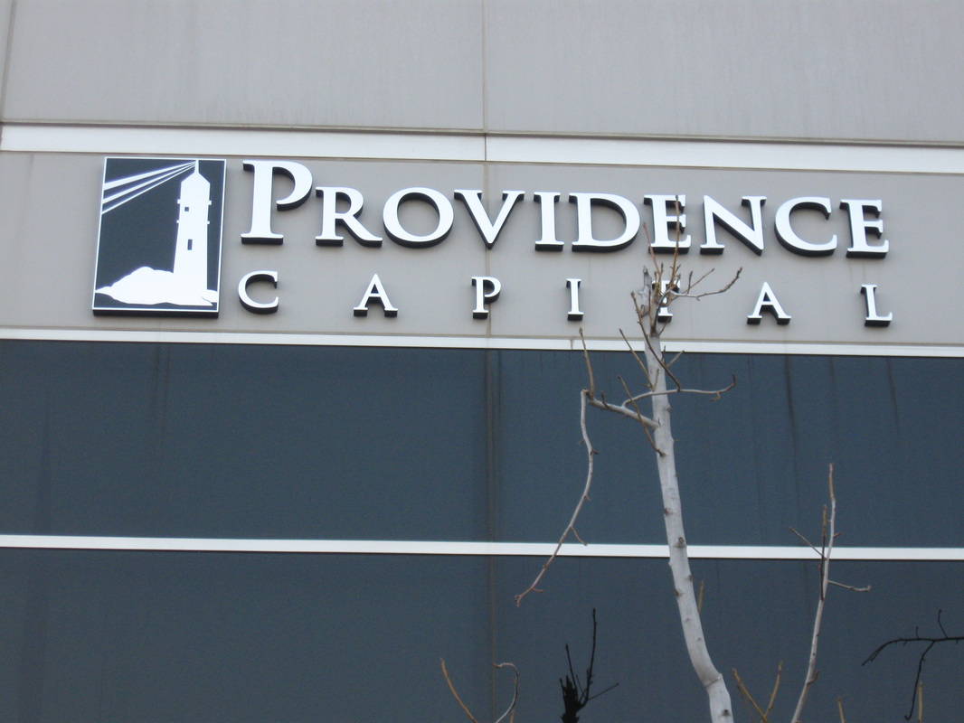 Exterior building fascia for Providence Capital: white dimensional letters spelling PROVIDENCE CAPITAL beside a black-and-white lighthouse logo panel, mounted on a gray precast band above dark glazing