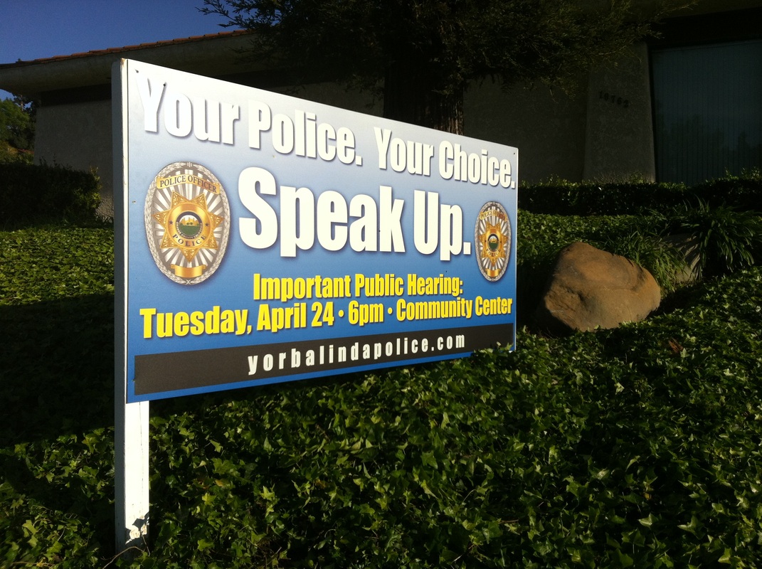 Post-mounted exterior sign on a landscaped slope reading 'Your Police. Your Choice. Speak Up.' with two police badges and public hearing details, for Yorba Linda Police
